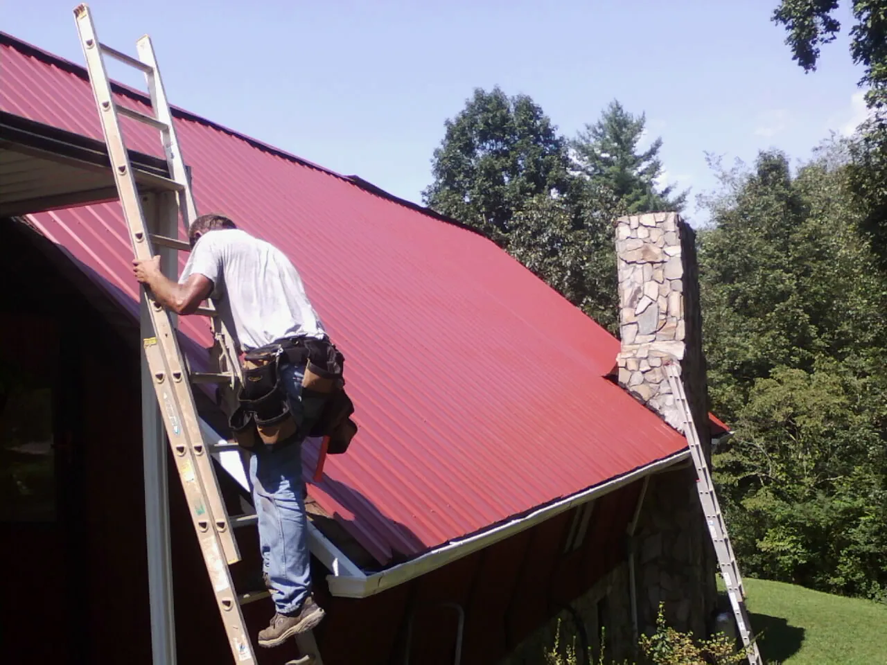 Roofer on a ladder beside a red metal roof and stone chimney — Soffit & Fascia Repair by Roofing Brothers in Tyngsboro